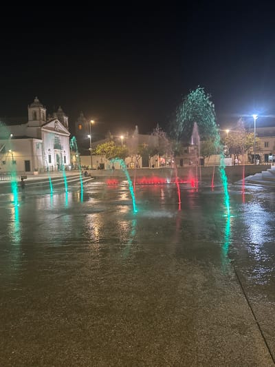 The image depicts a public square in Lagos, Portugal at night. A water fountain feature is prominently displayed in the foreground, with jets of water illuminated by green and red lights, creating a vibrant and dynamic visual effect. The water reflects on the wet pavement, adding to the atmospheric illumination. In the background, a white building with architectural features resembling a church or historic institution is visible, along with streetlights and trees. A few people are scattered throughout the scene, some appearing to be standing or sitting near the fountain or on steps leading up to the background structures. The overall scene suggests a lively urban environment at night, likely a popular gathering spot.