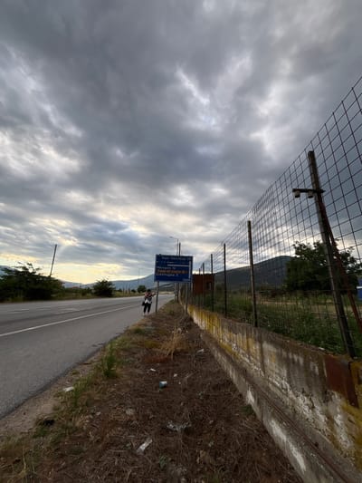 The image captures a roadside scene under a cloudy sky. A paved road runs from the left to the center of the frame, bordered by a grassy and somewhat littered verge. A person, a woman with dark hair, wearing a white top and dark bottoms, is standing on the side of the road, looking at their phone. A blue road sign is visible behind her, indicating distances to "Kato Nevrokoopi 25", "Mikropoli 12", "Cave of Angitis 5", and "Kokkinogeia 3". The sign also displays "150M". To the right, a wire fence runs along the scene, supported by wooden posts. Beyond the fence, there is overgrown vegetation and a distant view of hills or mountains. The overall environment suggests a rural or semi-rural area, likely in the Prosotsani Municipal Unit of Greece, given the Greek text on the road sign.