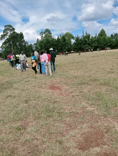 The scene depicts an outdoor grassy field in Lwandeti ward, Kenya, under a partially cloudy blue sky. In the mid-ground, a group of approximately 15-20 individuals, appearing as adults and young adults in casual attire, are gathered, predominantly facing towards the right, observing an activity. Some light-colored items, possibly bags or clothing, are visible on the ground near their feet on the left. Further in the background, a football (soccer) match is in progress on the same field, involving at least 8-10 players wearing red and green/light green jerseys. A white football is visible in mid-air, and goalposts are present on the right side of the playing area. The field surface consists of dry, short grass with visible patches of exposed soil. The background features tall, broadleaf trees on the far left and a dense line of coniferous trees bordering the playing field across the right and center.