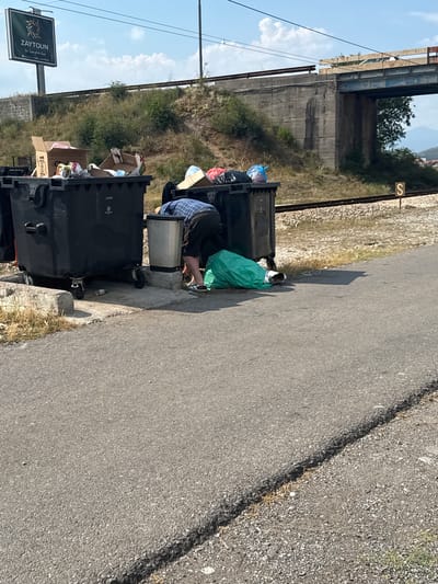 The image depicts an outdoor scene in Mojanovići, Montenegro, likely during the daytime with clear skies. The primary subjects are several large black trash bins overflowing with garbage. One person, dressed in dark clothing and a blue and white checkered shirt, is bent over, rummaging through one of the bins. A bright green garbage bag is also visible, partially in a bin and partially on the ground.

The setting appears to be along a roadside, with a paved surface to the right and a gravel embankment with some vegetation to the left. In the background, a concrete overpass or bridge supports is visible, with train tracks running parallel to the embankment. A small sign with the letter "S" is mounted on a post near the tracks. Further in the distance, hills and trees suggest a semi-rural or industrial area.

The overall impression is one of neglect and untidiness due to the overflowing trash bins. The activity of the person suggests they are searching through the refuse, possibly for recyclables or discarded items. The sunlight indicates it is daytime, and the clear sky suggests fair weather. The presence of the "ZAYTOUN" sign in the lower left corner is a notable detail, providing a potential clue to the location's commercial context. This sign features a logo with olive branches and the text "ZAYTOUN THE TASTE OF THE EAST".