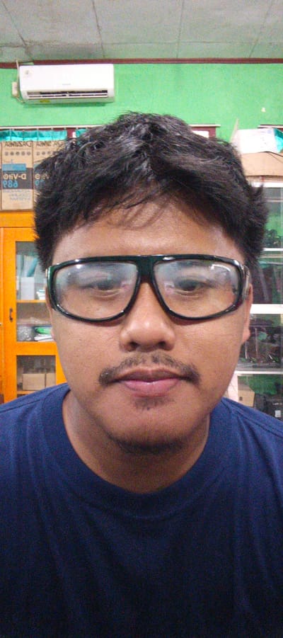 A medium shot captures a man from the chest up, positioned indoors. He wears thick, dark-rimmed glasses and a dark blue t-shirt. His hair is dark and tousled. He has a mustache and a small patch of stubble below his lower lip. The background is a bright green wall with a white air conditioning unit mounted on it, above a wooden trim. To the left, part of an orange cabinet is visible, and beyond that, shelves filled with various objects, including boxes and what appear to be electronics. To the right, shelves are also visible with a blurred background that suggests more shelving or displays. The lighting is moderate, creating some glare on his glasses. The image appears to be taken in Sukabumi, Indonesia, likely within an office or retail environment.