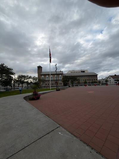 In Bodø, Norway, a vast, overcast public square is dominated by a red brick paved surface in the foreground. To the left, a curved concrete walkway leads towards a grassy area where a lone individual, appearing to be a child, walks away from the camera. A tall flagpole with a Norwegian flag stands prominently in the middle ground. Behind the flag, a large, light-colored building with numerous windows and a distinctive tower is visible, likely housing governmental or public facilities. Trees line the background, and other smaller buildings can be seen further in the distance. The sky is heavily clouded, indicating a possibly cool or rainy day.