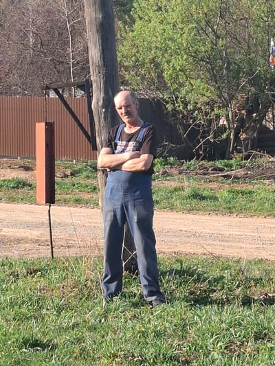 The image shows a man standing in an outdoor setting. He has a bald head and is wearing a black T-shirt underneath blue overalls. His arms are crossed. He is standing on a dirt and grass path. In the background, there is a building with a brown exterior and corrugated metal roofing. Additionally, there are trees and grass visible in the surrounding area. The location is possibly in Kamennyi Kliuch, Russia. A wooden post is visible to the left of the man.