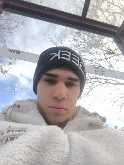 A male individual is depicted in a close-up image, situated within an outdoor shelter, likely a bus stop, in Freital, Germany. He wears a black knit beanie cap with white block letters spelling "GEEK" and a cream-colored, zippered fleece jacket featuring visible drawstrings. His facial expression is neutral as he looks directly at the camera. The background, viewed through transparent panels of the shelter, shows a partly cloudy sky and deciduous tree branches largely devoid of foliage, indicative of autumn or winter. On a transparent panel to the upper left of the individual, a hand-drawn swastika symbol is visible. Further to the right on a reflective surface within the shelter structure, reversed text "4QZ" is discernible.