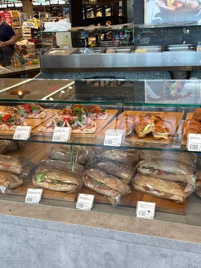 The image shows a refrigerated display case filled with various food items in a retail establishment. In the foreground, there are several wrapped sandwiches, some appearing to be baguettes with fillings like lettuce, tomato, and cured meat. Behind these, on wooden boards, are portions of pizza topped with vegetables and meat, as well as croissants with cheese and meat fillings. In the background, a person wearing a dark shirt is partially visible to the left. The establishment appears to be a food service area, possibly in a supermarket or deli, with shelves and display units stocked with products. The setting is brightly lit, and the overall impression is one of a food retail environment offering prepared meals. There is no discernible information within the image to indicate the location is Mojanovići, Montenegro.