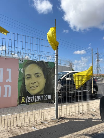 The image displays an outdoor scene in Kfar Yona, Israel, featuring a wire mesh fence with attached banners and flags under a partly cloudy blue sky.
A large banner is affixed to the fence, prominently featuring a portrait of a smiling woman with dark, curly hair, identified by Hebrew text as "ענבר הימן (27)" (Inbar Haiman (27)), accompanied by a yellow ribbon symbol. The banner has a green and pink background. Another partial banner, primarily pink with Hebrew text, is visible to the left.
Several bright yellow flags, made of fabric, are attached to the fence posts with wooden sticks, visible on both sides of the banner and further down the fence line. The metal fence is topped with barbed wire.
Behind the fence, a dark-colored SUV is partially visible, parked or driving on a paved surface. Further in the background, a large metal lattice power transmission tower stands against the sky, with power lines running horizontally across the frame. The ground directly in front of the fence shows debris, including paper and other small items.