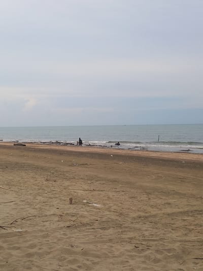 A wide, slightly overcast view of a sandy beach in Gampongkuala, Indonesia. The sand is a light brown color and shows some debris like scattered plastic and driftwood. The ocean is visible in the background, with gentle waves lapping at the shore. The water is a muted green-blue. On the beach, near the water's edge, there are a few scattered pieces of driftwood and some scattered litter. Two indistinct figures are present on the shoreline: one, appearing darker and seated, is closer to the water, while another, lighter figure, is further down the beach and also near the water. A single dark vertical pole stands in the water. The sky is a pale gray with hints of white clouds.