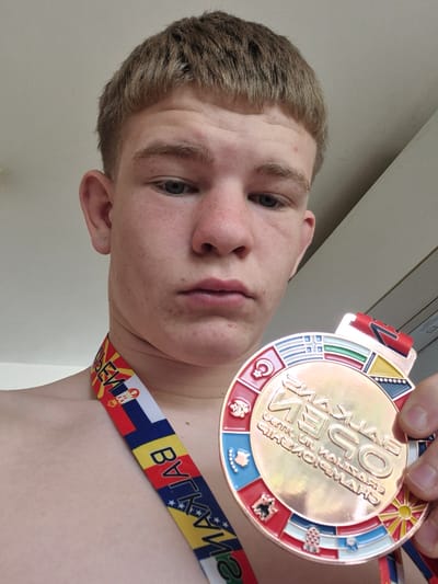 A young person, likely male, with light brown hair and fair skin, is depicted in a close-up shot, looking directly at the camera. The subject is holding up a large, round, bronze-colored medal with their right hand, positioning it prominently in the foreground.
The medal features a central embossed design with Cyrillic text. Clearly visible are "БАЛКАНСКО" (Balkan), "2023", and "МАКЕДОНСКИ ШАМПИОНАТ" (Macedonian Championship), indicating an event related to the Macedonian Championship in 2023, possibly a Balkan-wide competition. The medal's border is adorned with various national flags or emblems in colorful enamel, including the sun from the Macedonian flag, a checkerboard pattern reminiscent of the Croatian flag, a double-headed eagle possibly from the Albanian flag, a crescent and star similar to the Turkish flag, and symbols from the Bosnian flag.
The medal is suspended from a multi-colored ribbon draped over the person's left shoulder. This ribbon also features national flags, specifically the Macedonian, Serbian, and Bulgarian flags, along with Cyrillic text such as "МАКЕДОНСКИ" (Macedonian) and "БЪЛГАРИЯ" (Bulgaria), reinforcing the regional Balkan context of the event.
The background is a simple, light-colored surface, possibly an interior wall or ceiling, suggesting an indoor setting. The photo is taken from a slightly low angle, with the subject's head tilted back. The person has a neutral to slightly content expression, proudly showcasing their medal. The scene captures a moment of achievement from what appears to be a regional championship, aligning with the provided context of Beloslav, Bulgaria, as a location in the Balkan region.