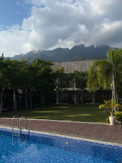 The image depicts a tranquil resort setting in Pasir Putih, Indonesia, characterized by a swimming pool, lush greenery, and a backdrop of majestic mountains. In the foreground, a vibrant blue swimming pool with tiled edges and a wooden deck area is visible. A set of stainless steel pool ladders extends into the water, reflecting on its surface. Beyond the pool, a well-maintained lawn stretches towards a two-story building with a tiled roof, featuring multiple balconies and windows. Mature trees and palm trees are interspersed throughout the property, adding to the tropical ambiance. In the distance, a mountain range is partially shrouded in clouds, creating a dramatic and serene landscape. The scene is devoid of any people, emphasizing the peaceful and secluded nature of the location. The lighting suggests it is daytime, with the sun casting shadows on the lawn.