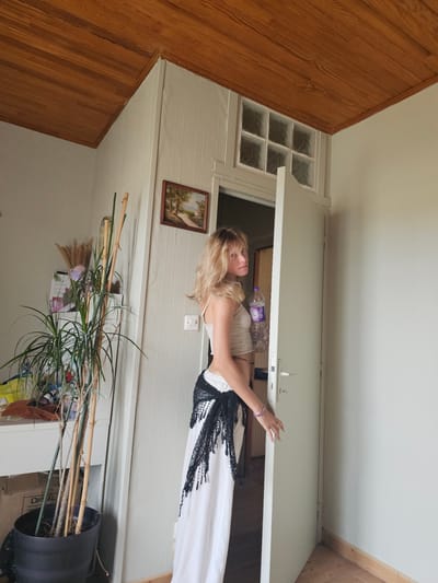 A blonde woman stands in an open doorway. She is wearing a white dress with a black decorative overlay at the waist. She is holding onto the doorframe with her right hand and her left arm is bent, with her hand resting on her hip. She has a bottle of water on a shelf behind her. The room is furnished with wooden floors and walls. There is a small framed painting on the wall to the left of the doorway. A tall potted plant is in the foreground. The image appears to be taken during the daytime, likely indoors in a residential setting.