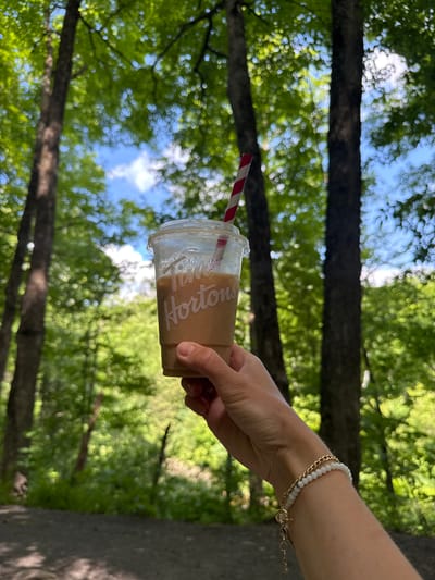 In Chelsea, Canada, a person's hand holds a clear plastic cup with a red and white striped straw, containing a light brown beverage. The cup features a white logo that reads "Tim Hortons." The hand is adorned with a bracelet made of small white beads and gold chain. The background is a lush forest with trees and green foliage, suggesting an outdoor setting. The sunlight filters through the leaves, creating a dappled effect. The ground in the foreground appears to be a gravel path.