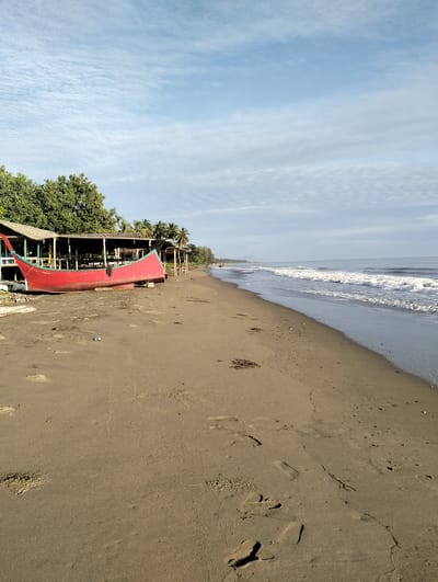 The image depicts a beach scene in Sagoe, Indonesia. A red boat rests on the sandy shore near a wooden structure with a thatched roof and wooden supports. The beach appears to be made of dark sand and has footprints leading towards the water. The sea has gentle waves rolling towards the shore. The sky is light blue with wispy clouds. The trees and vegetation are present along the coastline.