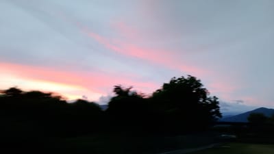 The image captures a blurred sunset scene over Gorno Sahrane, Bulgaria. The sky is dominated by soft pink and white clouds streaked across a pale blue canvas. Silhouetted against the vibrant sky are dark, indistinct shapes of trees forming a line along the horizon. In the distance, the faint outlines of mountains are visible, partially obscured by the low light. The overall impression is one of motion blur, suggesting the photograph was taken from a moving vehicle.