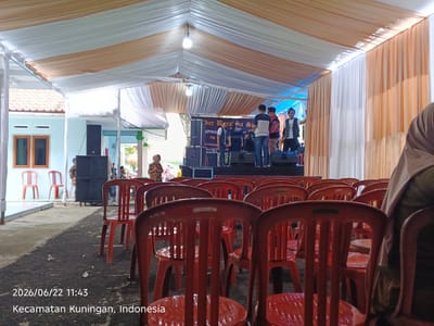 An outdoor event setup is depicted under a large tent with white and tan fabric. In the background, a stage is set up with a banner displaying Indonesian text that appears to be an event title and contact information. Several individuals are standing on the stage, some appearing to be performers or speakers. In the foreground, rows of bright red plastic chairs are arranged, facing the stage. The ground appears to be a paved or concrete surface, with some debris scattered around. A watermark in the bottom left corner indicates the date and location as "2026/06/22 11:43 Kecamatan Kuningan, Indonesia". To the left of the stage, there are some speakers and what appears to be a building with orange-framed windows.