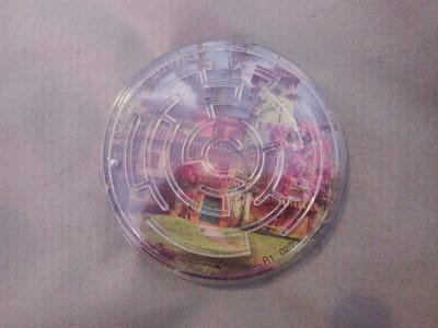 The image shows a circular plastic maze with a Disney-themed image of a house and lush vegetation. The maze is transparent, allowing the image to be seen underneath. The text "©Disney" is visible on the left side of the maze. On the bottom right of the maze, there is a date stamp that reads "R1 02/23". The maze is placed on a textured, grey surface, possibly a fabric or a mat. There is no indication of the city of Inhulo-Kamianka, Ukraine, in the image.