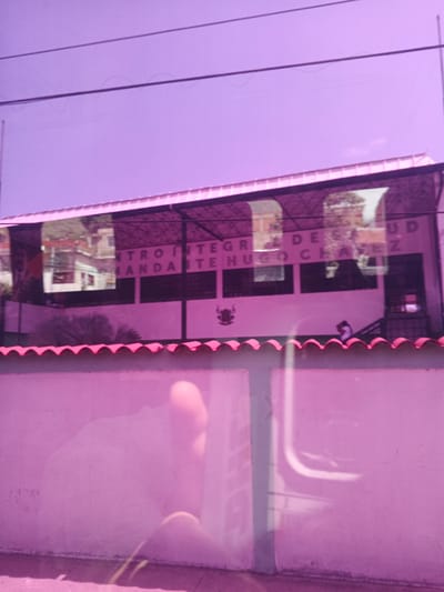 The image shows the exterior of a building with text visible on its facade. The building appears to be a health center, as suggested by the text "CENTRO INTEGRAL DE SALUD HUGO CHAVEZ" which is partially visible. The building has a white roof and a large glass facade. Through the glass, reflections of surrounding buildings and trees are visible. There are also dark rectangular panels on the facade, possibly windows or signs. A small crest or emblem is visible on the wall below the text. In front of the building, a lower structure with a pink, wavy roof is present, which might be a wall or another part of the facility.
Two figures are partially visible near the right side of the building, one appearing to be walking up a set of stairs. The overall lighting suggests it is daytime, with bright sunlight casting reflections. The image has a strong purple tint, which may be due to lighting conditions or a filter applied to the photograph. The scene is likely outdoors in a populated area, given the presence of buildings and power lines overhead. The wavy pink roof in the foreground adds a distinctive visual element to the scene. The location context is Macuto, Venezuela, which aligns with the Spanish text on the building.