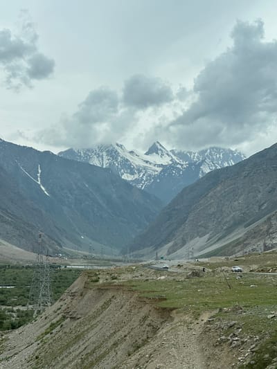 An outdoor, eye-level, vertical shot captures a vast mountain landscape under a cloudy sky in Pandrass, India. The image is dominated by imposing, snow-capped mountains in the background, their peaks obscured by clouds. The slopes of the mountains are a mix of rocky terrain and patches of snow. In the mid-ground, a road winds through the valley, with a few vehicles visible. To the left, a lush green valley is dotted with trees and a body of water. Several tall, metallic electricity pylons are scattered across the landscape, indicating infrastructure. The foreground features a steep, rocky slope with sparse vegetation. The overall impression is one of natural grandeur and the juxtaposition of human infrastructure within a rugged environment.