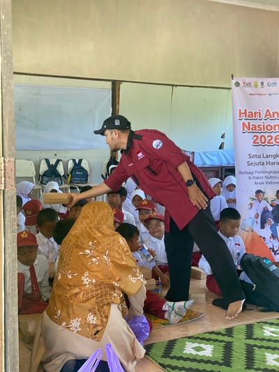 In a school classroom in Handil Baru, Indonesia, a man wearing a maroon shirt, black pants, and a black baseball cap is handing a small, light brown box to a group of children. The children are seated on the floor, many wearing white shirts and some with red hats. The man's shirt has patches on the sleeves and chest. Behind the children, there are several white chairs with blue backpacks on them. A banner in the background has Indonesian text and an image of children. The floor is covered with a green and white patterned rug. The room appears to be rustic, with a textured wall.
