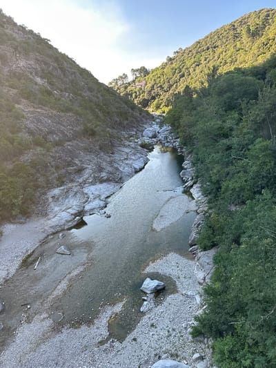 The image depicts a scenic view of a river flowing through a valley. The riverbanks are lined with rocks and trees, with the terrain rising into tree-covered hills on either side. The water is clear and reflects the light, with some areas of shallow riverbed visible. The scene appears tranquil and natural, with no human presence or activity evident. The lighting suggests it is either early morning or late afternoon, with the sun casting shadows and illuminating parts of the landscape. Based on the provided context, this image is likely of a location in Générargues, France.
