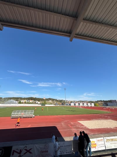 The image depicts a soccer field and running track in Valpaços, Portugal, adjacent to commercial banners and buildings. The sky is blue with wispy clouds. People are present near the banners, while soccer players are on the field, one near a sheltered area and another running. Sprinklers are watering the field. Buildings and trees dot the background landscape.