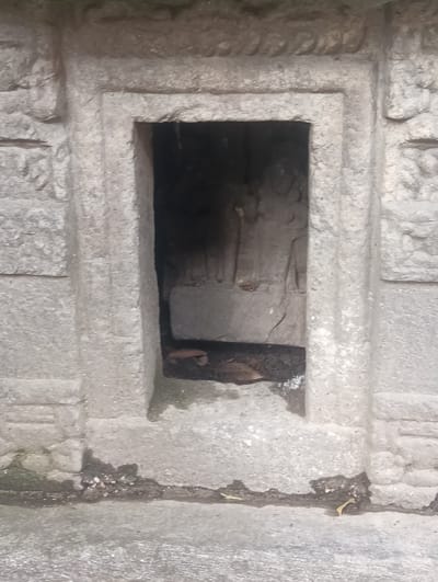 This is a close-up, eye-level, full outdoor shot of a recessed stone niche carved into a stone wall. The niche itself is dark, with some lighter-colored debris and a few dry leaves visible at the bottom. Within the niche, a carved relief is dimly visible, appearing to depict two standing figures. The stone surrounding the niche is rough-hewn and appears to be weathered. The wall has decorative elements on either side of the niche, with horizontal lines and some textured patterns. The ground in front of the wall is also stony and appears to have some damp patches and debris. The image is not in focus. There is no indication that this location is in Gojra, India.