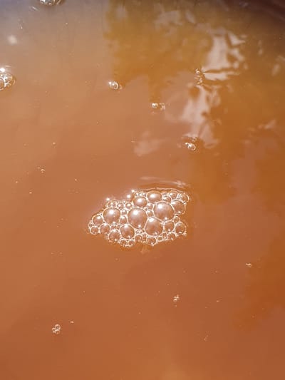 This is a close-up, top-down, full-frame shot of a shallow body of murky, brown liquid with a concentration of frothy bubbles on the surface. The liquid appears to be water with suspended sediment. The bubbles are varied in size, with a prominent cluster of larger bubbles in the lower-middle section of the image, reflecting light from above. Smaller, isolated bubbles are scattered throughout the liquid. The surface has a slight sheen, indicating it is a liquid. There are no people, animals, or discernible structures visible. The image is a still shot of a static liquid surface. There is no information in this image to determine its location in the city of Novoivanivka, Ukraine.