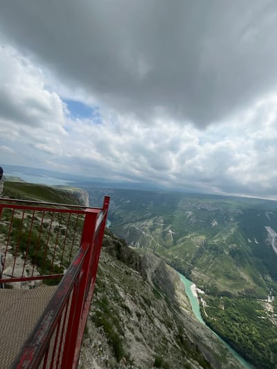The image presents a high-angle view from a location with a red metal safety railing. The landscape includes mountains and a body of water. The foreground is dominated by the railing structure. Beyond the railing, the terrain slopes steeply down into a valley with varying vegetation. A river or lake winds through the valley floor. Clouds cover a significant portion of the sky. The location is likely in the vicinity of Dubki, Russia.