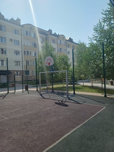 This media file captures an outdoor scene on a sunny day, likely in Elitnyi, Russia. The image is oriented vertically, showcasing a sports court with a basketball hoop and a small soccer goal. The court surface appears to be a dark red, with a contrasting green section marked by a white line. A metal fence surrounds the court, separating it from a paved area and a grassy strip with trees.
In the background, a light-colored brick apartment building with numerous windows rises several stories. The building has an unusual curved architectural design and appears to be residential. Lush green trees with dense foliage are visible, partially obscuring the building and suggesting a pleasant, warm-weather environment.
The time of day is indicated by the bright sunlight and the long shadows cast by the fence and the soccer goal, suggesting it is either morning or late afternoon. The sky is clear and blue, indicating fair weather. There are no people visible in the scene, and no text is present. The overall impression is one of a quiet, everyday scene on a recreational area next to a residential building.