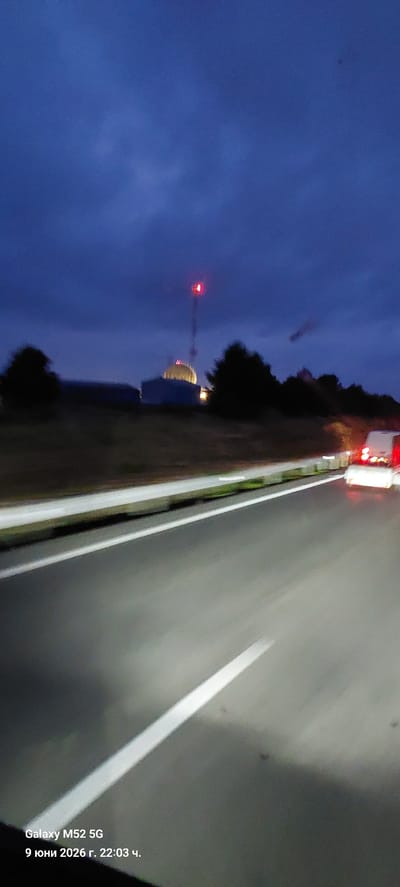 The image depicts a nighttime view from a moving vehicle on a highway, likely in or near Vandellòs i l'Hospitalet de l'Infant, Spain. The sky is dark blue, with some lighter clouds. In the distance, a radio tower with a red light at its top is visible, along with a domed structure, possibly part of a facility or building. Silhouetted trees line the side of the road. The highway itself is paved with white lane markings. Two vehicles with illuminated taillights are ahead on the right lane, suggesting traffic is moving. The bottom left corner of the image contains text indicating "Galaxy M52 5G" and a date and time: "9 юни 2026 г. 22:03 ч.".