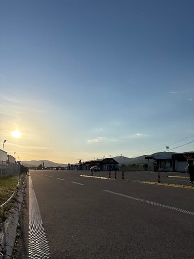 The image depicts an asphalt road in Dimitrovgrad Municipality, Serbia, running adjacent to buildings and a mountain range. The sky transitions from blue to a golden hue near the horizon, where the sun is positioned. A concrete barrier separates the road from an adjacent grassy area. The road features a painted white line and tactile paving. Cars are parked along the side of the road near the buildings. Wires run parallel to the road above the buildings. A person is visible in the top corner of the image.