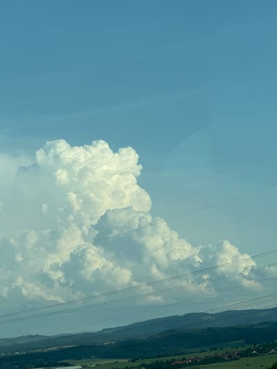 A wide-angle, vertical photograph captures a dramatic sky over a rural landscape, likely in the municipality of Hörselberg-Hainich, Germany. The upper two-thirds of the image are dominated by a brilliant blue sky, interrupted by a colossal cumulus cloud formation that rises prominently from the horizon. This cloud is voluminous and textured, with bright white highlights indicating direct sunlight and darker, shadowed undersides suggesting depth and mass. Faint, wispy cirrus clouds are subtly visible in the upper portions of the sky.

Stretching across the frame, from left to right, are multiple parallel power lines, suspended by unseen poles. These lines are thin and dark, creating a linear pattern that contrasts with the organic shapes of the clouds and landscape.

The lower third of the image depicts a rolling, verdant landscape. Dark green, forested hills form a backdrop, with visible lines of trees. In the middle ground and foreground, patches of lighter green fields are interspersed with small clusters of red-roofed buildings, suggesting a village or scattered rural dwellings. The overall impression is one of a vast sky above a serene, cultivated terrain. The lighting suggests it is daytime, possibly late afternoon, given the contrast and shadows.