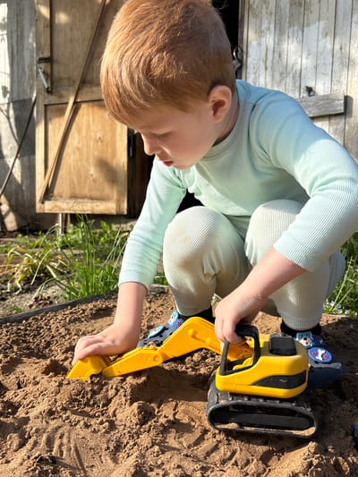 A young child in light green ribbed clothing is playing with a yellow toy excavator in a sandbox. The child is squatting, manipulating the excavator's arm in the sand. The sandbox is edged with a dark border and contains brown sand. To the left, there is a weathered wooden structure, possibly a shed or a building, with visible hinges and a latch. The location of the scene is not definitively discernible as being in the city of Италмас, Russia from the provided image details.