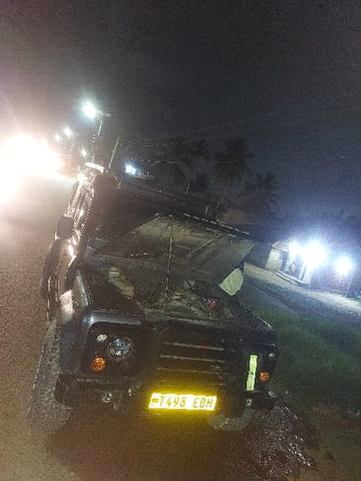 This nighttime image depicts a dark-colored off-road vehicle, specifically resembling a Land Rover Defender, with its hood raised, indicating an open engine compartment. The vehicle is positioned on the unpaved shoulder of a road in Msata, Tanzania. Its front features round headlights, orange turn signal indicators, and a black grille. A bright yellow Tanzanian license plate, "T498 EBH," is affixed to the front bumper. The ground immediately surrounding the vehicle, especially on the right, is muddy and saturated, with puddles reflecting light. To the left, several bright streetlights illuminate the road extending into the distance. On the right side of the frame, silhouetted palm trees are visible against the dark sky, and a distant structure emits a strong, bright light. No individuals are visible within the frame, and no specific actions or interactions are depicted beyond the vehicle's open hood.