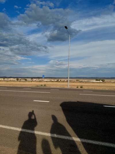 A streetlamp stands tall on the right side of a two-lane asphalt road, casting a long shadow across the pavement. In the foreground, two distinct human shadows are visible, one of which is holding what appears to be a smartphone, suggesting a selfie is being taken. Beyond the road, a dry, grassy plain stretches out, dotted with small buildings that form a rural settlement. In the distance, rolling hills are visible under a dramatic sky filled with a mix of blue patches and heavy, grey clouds. A blue traffic sign with a white symbol is planted on the side of the road. The overall scene depicts a roadside view in a semi-arid region of Kazakhstan, possibly within or near Қапанбұлақ ауылдық округі.