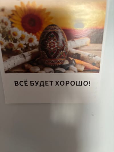 A decorative Easter egg rests among daisies and a sunflower. The egg is ornate with intricate red, green, and gold patterns. Behind the egg, a vibrant yellow and orange sunset or sunrise is visible. Several smooth, gray stones and a piece of birch bark lie on a wooden surface in the foreground. Below the image, the Russian phrase "ВСЁ БУДЕТ ХОРОШО!" is printed in black, capital letters. This phrase translates to "Everything will be fine!". This image likely originates from a postcard or decorative item associated with Easter traditions in Russia, possibly in the city of Imeni Morozova, given the context of Russian language and cultural elements.