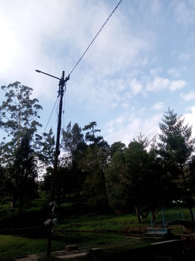 The image shows a landscape in Mya Sit Kaing Ywar Thit, Myanmar, featuring a green area with trees and a small pond. A utility pole with a streetlight stands prominently in the foreground. A small blue bridge crosses part of the pond. The sky is partly cloudy.