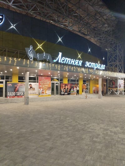 The image shows the facade of a building with the Russian text "Летняя эстрада" (Summer Stage) illuminated in large blue letters across the top. Musical notes and symbols are also part of the building's signage. The building has a covered entryway with multiple glass doors and is decorated with strings of small lights. Posters are displayed on the exterior walls advertising various events and performers, including "RADIO TAROK", "КУБАНСКИЙ КАЗАЧИЙ", and "ХАСМИ". There are several people visible in the foreground and near the entrance. One person in a grey shirt and shorts is walking away from the camera on the paved plaza in front of the building. Two people stand near a poster display, and another person in a light-colored shirt and shorts is standing near an entrance. The environment appears to be an outdoor entertainment venue at night, with artificial lighting illuminating the scene. The paving stones in the foreground are arranged in a geometric pattern. The city of Vinogradny, Russia, is mentioned in the prompt, but this specific location is not visually identifiable from the image alone without further context.