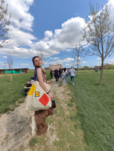The image captures a sunny day at an outdoor location in Neofit Rilski, Bulgaria. The sky is blue with puffy white clouds. A dirt and gravel path leads towards a historical building in the distance where a group of people are walking. To the left of the path, there's a grassy area with a small wooden structure resembling a shed or barn.

A young woman is the focal point of the image, standing on the path and looking at the camera. She is carrying a large tote bag with the Lidl logo on her shoulder. She is dressed in a casual outfit, and holds a bottle in her hand. A child sits in a stroller near the wooden shed to the left. Other people are walking along the path towards the building. A few trees are scattered throughout the green grassy area.