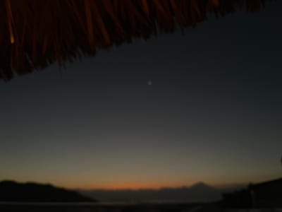 The image captures a twilight scene over a body of water, likely the ocean given the coastal silhouette of landmasses. The sky transitions from a dark, dusky blue near the top to lighter shades of gray and orange near the horizon, indicating either sunset or sunrise. A blurred, distant mountain range is visible against the horizon. In the foreground, the edge of a thatched roof or awning, made of dried plant material, partially obscures the top portion of the image. A small, circular, bright object, possibly the moon or a bright star, is visible in the upper-middle section of the dark sky. The overall scene is dark and slightly out of focus, creating a moody atmosphere. The location is Kertasari, Indonesia.