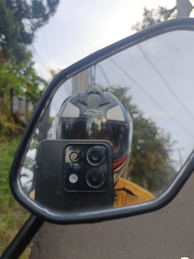 The image depicts a unique selfie captured indirectly through a motorcycle's rearview mirror in Sindangasih, Indonesia. The primary subject is a black smartphone, held up to the mirror, effectively taking a picture of its own reflection. The phone's rear camera module is distinctly visible, featuring multiple lenses and a flash. Text on the phone's back identifies it with "9M80" and "MACRO LENS".

Reflected within the mirror, behind the smartphone, is a person wearing a black and white motorcycle helmet. A prominent "S" logo is visible on the back of the helmet, which also features white triangular accents. A portion of a yellow garment, likely a shirt or jacket, is visible on the wearer's shoulder.

The surrounding environment, both directly visible around the mirror's frame and reflected within it, shows an outdoor setting during daytime. Lush green trees and foliage line what appears to be a roadside, suggesting a natural or semi-rural area. The sky above is bright but largely overcast, indicating cloudy weather. A road surface is subtly discernible in the reflection below the helmet, reinforcing the context of a motorcycle journey. The activity captures a moment of rest or a quick photo stop on the road.