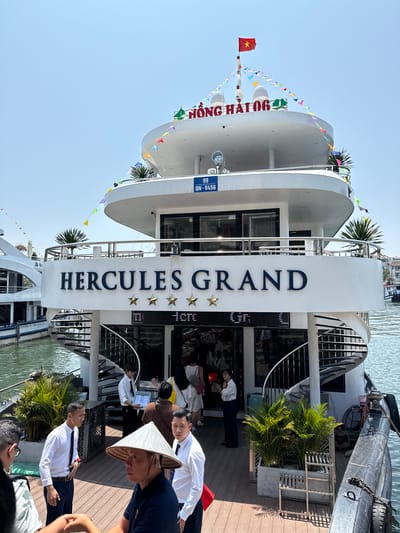 This media file captures a sunny daytime scene at a pier or dock in Tuan Chau Ward, Vietnam. The main subject is a large, multi-deck white boat or cruise ship, adorned with colorful triangular bunting flags, suggesting a festive or tourist setting.

Prominently displayed on the boat's bow is the name "HỒNG HẢI 06" in red letters, with a registration number "99 QN-9456" visible below it. A large, vertical white structure on the side of the boat clearly shows the name "HERCULES GRAND" in dark blue capital letters, accompanied by three gold stars, indicating a rating or class. A small Vietnamese flag (red with a yellow star) flies from a mast on the boat's top deck.

Several people are visible on the wooden deck beside the boat. Most appear to be staff or crew, dressed in white collared shirts and dark trousers, possibly guiding passengers. One person in the foreground wears a traditional Vietnamese conical hat (nón lá), and another is holding a small Vietnamese flag. Other individuals can be seen seated or standing inside the boat's lower deck, suggesting a boarding or preparation for departure. Lush green palm plants in large planters decorate the deck area. The water next to the boat is calm, reflecting the bright sky. The overall scene depicts activity around a tourist cruise or tour boat.