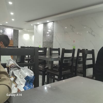 A photograph taken from an interior perspective shows a stack of dark-colored wooden chairs stored in a room. The location appears to be a commercial establishment or restaurant, suggested by the tiled floor, recessed lighting in the ceiling, and table. A person is partially visible in the lower part of the frame. The "Galaxy A14" watermark indicates the device used to capture the image. The exact location within the city of Nuseirat, Palestinian Territories, is not discernible from the visual elements in the photograph.