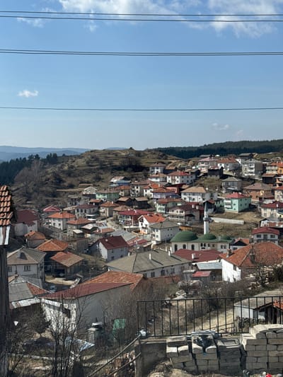 The image depicts an expansive elevated view of a hillside village identified as Kasak, Bulgaria, under a clear blue sky with sparse white clouds. In the upper portion, four dark horizontal power lines traverse the frame. The foreground, at the bottom, includes a partial view of a terracotta-tiled roof on the far left, a black metal fence on the right, and a stack of light-colored, rectangular construction blocks or stones below the fence. Bare deciduous tree branches are visible around the foreground elements.
The midground is dominated by a densely packed collection of multi-story residential buildings, varying in architectural style and color. Common house colors include white, grey, light blue, mint green, red, pink, and orange. The roofs are predominantly covered with red or terracotta tiles, with some featuring darker grey or brown materials. A distinctive mosque is centrally located within the village, identifiable by its large, multiple green domes and a tall, slender white minaret topped with a dark, conical cap. Several houses show clotheslines with hanging laundry.
The background consists of rolling hills and mountainous terrain. Some hills are sparsely covered with dry, light brown vegetation, while others are densely forested with dark green coniferous trees. A faint, hazy horizon line separates the distant landscape from the sky. No individuals or direct interactions are visible.