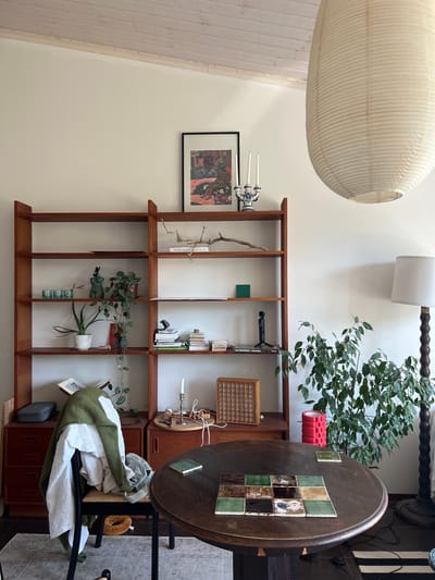 The image depicts an indoor scene, likely a living room or dining area. Dominating the center is a dark, round wooden table with a grid of decorative tiles arranged on its surface. To the left of the table, a tall, wooden bookshelf unit is filled with various items, including books, plants, and decorative objects. A chair is placed in front of the bookshelf, draped with a green sweater and a white garment. To the right of the table, a large potted green plant stands near a floor lamp with a white shade. A large, off-white paper lantern hangs from the ceiling in the upper right portion of the image. The room features white walls and a sloped, light-colored wooden ceiling. The overall impression is one of a lived-in, somewhat eclectic space. There are no people visible in the image. The location is identified as being in Hämeenlinna, Finland.