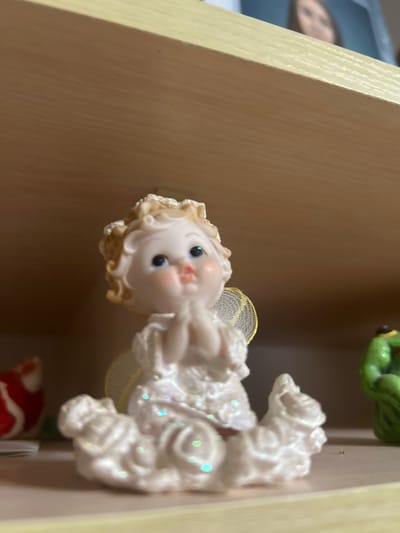 A small, ceramic or resin figurine of a light-skinned angel with blond, glitter-dusted curly hair, blue eyes, and rosy cheeks is centrally positioned on a light-colored wooden shelf in Dmitriyevskaya, Russia. The angel figurine wears a white, glitter-speckled gown with textured details and has translucent, mesh-like wings with yellow edging. Its hands are clasped together in front of its chest, and it sits on a white, cloud-shaped base. To the immediate left of the angel, a red and white object is partially visible and out of focus. To its right, an out-of-focus green figurine, possibly depicting a frog, is visible. Above the shelf and the figurine, a second, identical light-colored wooden shelf is present. In the upper right background, a blurred image of a person's face, possibly a young woman with dark hair, is visible on or behind the upper shelf. The scene is brightly lit, indicating an indoor setting.