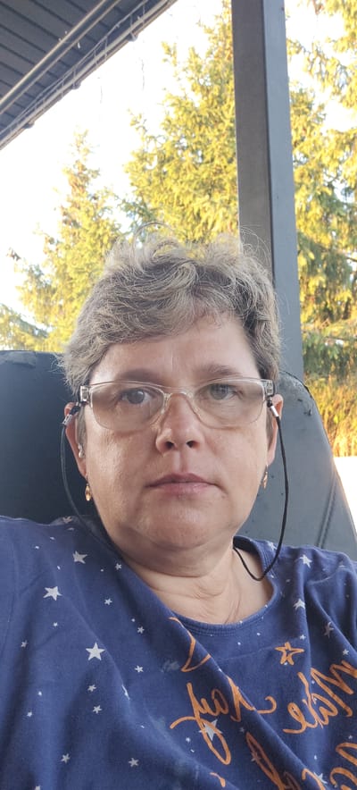This is a close-up, waist-up selfie of a middle-aged woman with short, graying hair and glasses. She is wearing a dark blue t-shirt with white stars and some orange text, the full text is not visible. The woman is wearing earrings and has a cord from her glasses draped around her neck. She is looking directly at the camera with a neutral expression.
The setting appears to be outdoors, possibly on a porch or patio, with a dark-colored chair behind her. In the background, there are green trees and a bright sky, suggesting it is daytime with clear weather. The lighting indicates it might be late afternoon or early evening, given the warm, golden tones on the trees.
There is no specific activity or event taking place; it's a posed portrait. No discernible emotions are evident beyond neutrality. The visible text on her shirt appears to be in a cursive font and is primarily orange in color, but it is not fully legible.