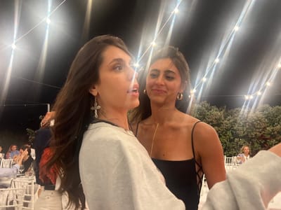 The image captures an outdoor nighttime event, possibly a wedding reception, with string lights overhead and lush greenery in the background. The focal point is two young women in the foreground, appearing to be in mid-conversation. The woman on the left, with long dark hair, is wearing a light-colored top and a large cross-shaped earring. The woman on the right, with dark hair pulled back and wearing a black dress, sports large hoop earrings and a delicate gold necklace. Their faces are close, suggesting an intimate moment. In the background, other guests are visible seated at tables, some of whom are indistinct. The overall ambiance is festive and social. The event is taking place in Fasano, Italy.