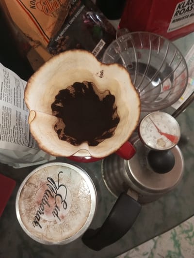 The image shows a coffee preparation scene with various elements arranged on a glass surface. A coffee filter with used grounds sits atop a red mug and a gooseneck kettle, which has a thermometer attached. Nearby is a clear plastic pour-over dripper.  A tin container with the words "Eid Mubarak" in stylized writing is on the left side of the image. Coffee bean bags are in the background, one with "Ellen Coffee" and another with "Klasik Roast Robusta." The image is shot from an overhead perspective.