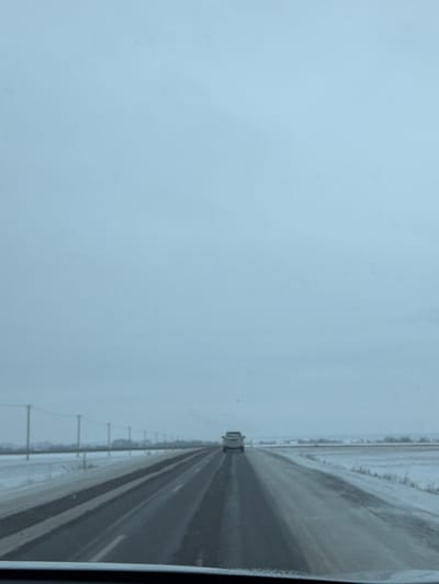 The image presents a forward-looking perspective from inside a moving vehicle on a multi-lane highway during winter in the city of Нижегородка, Russia. The road surface, appearing dark grey, is wet or slushy with visible tire tracks, indicating recent precipitation or melting. Faint lane markings are discernible on the asphalt. Snow covers the ground on both sides of the highway, extending into flat, open fields that reach the horizon. A line of utility poles, connected by horizontal wires, runs parallel to the road on the left side, receding into the distance. The sky overhead is uniformly overcast and light grey, suggesting a dull, possibly cold, day. In the distance, centered in the same lane of travel, a single light-colored SUV or pickup truck is visible, moving forward. No individuals are present in the scene. The bottom edge of the image reveals the dark dashboard or windshield frame of the viewpoint vehicle.