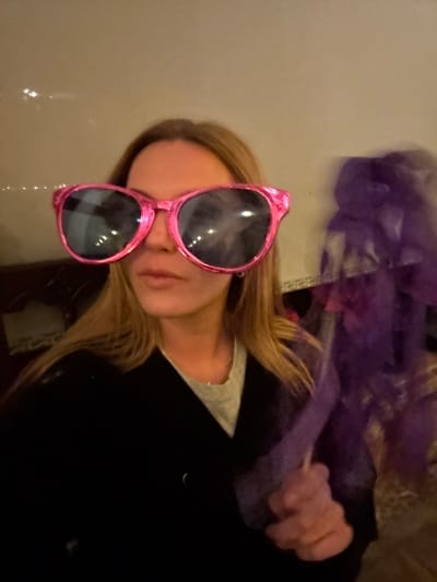 A person is seen wearing oversized pink glasses. They have blond hair and are wearing a black shirt with a light-colored shirt underneath. A hand can be seen holding purple fabric in the upper right corner of the frame. The background appears to be a plain light-colored wall. The image is slightly blurry and rotated.